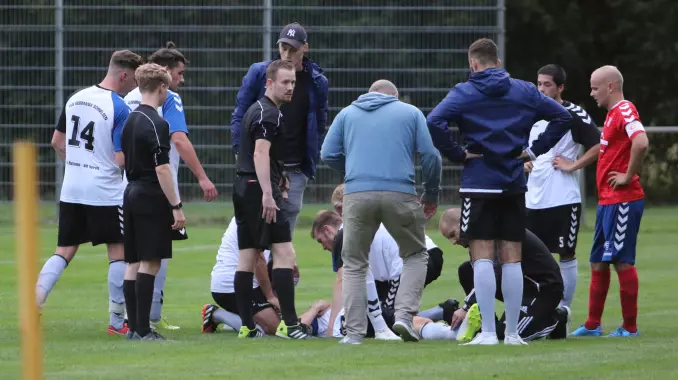 Spieler beider Teams, Referee Myles Taylor Tardieu (2. von links) und Schiedsrichter-Assistent Patrick Silvester (4. von links) stehen um den verletzt am Boden liegenden Schnelsener Dennis Swierkot herum.